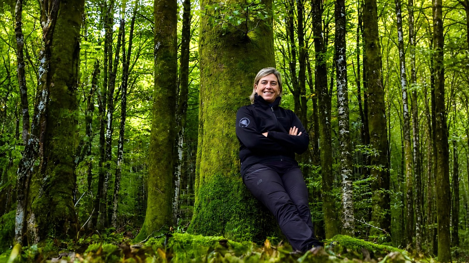 Camilla Costa nel bosco, sorridente, appoggiata a un grande albero coperto di muschio in una foresta verde.